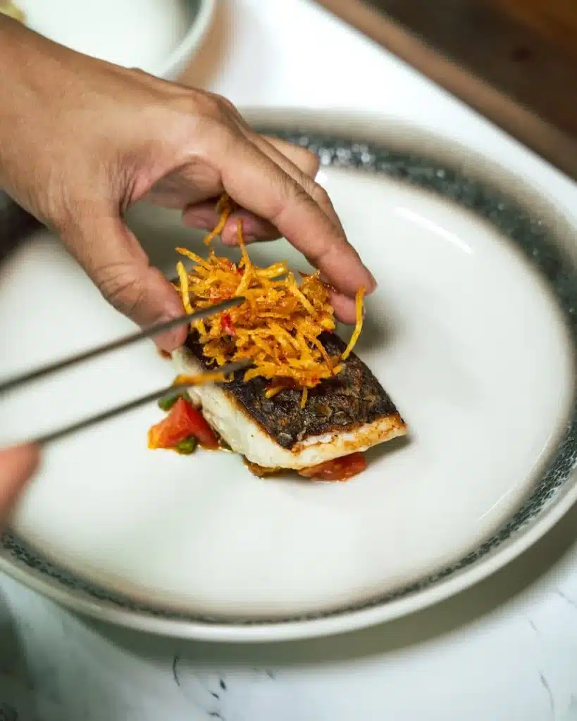 Chef plating a seared fish fillet with crispy garnish, an onboard fine dining dish on Otium